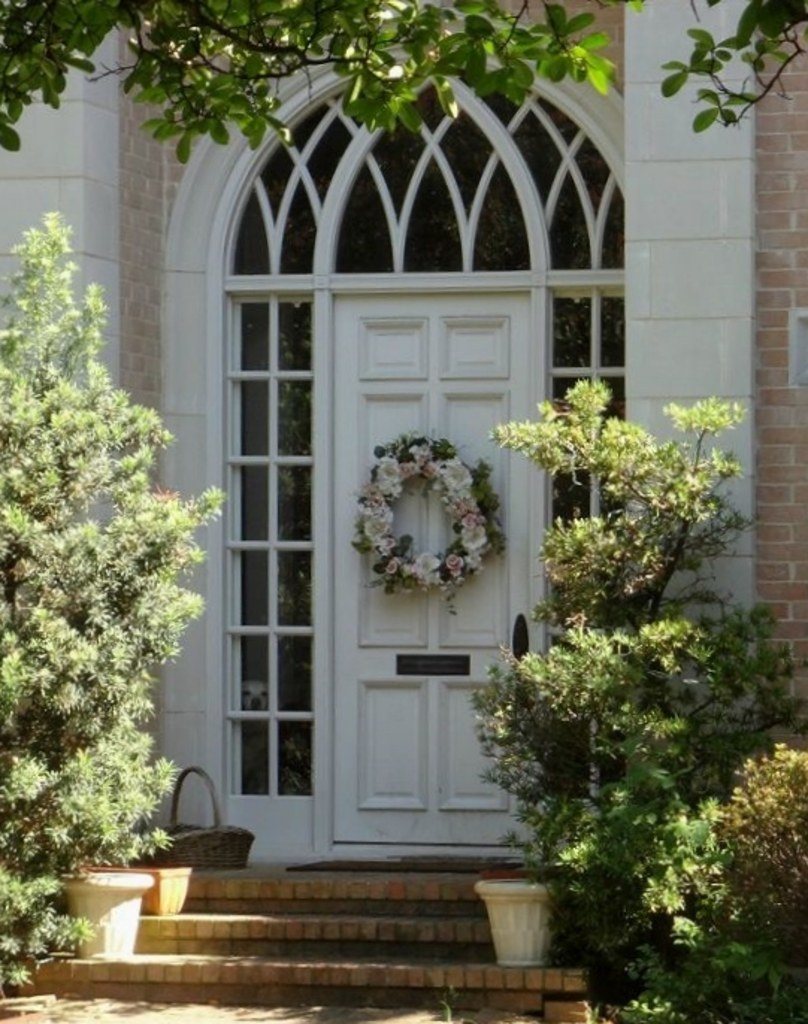 White front door with an arched fanlight and a wreath between potted evergreens